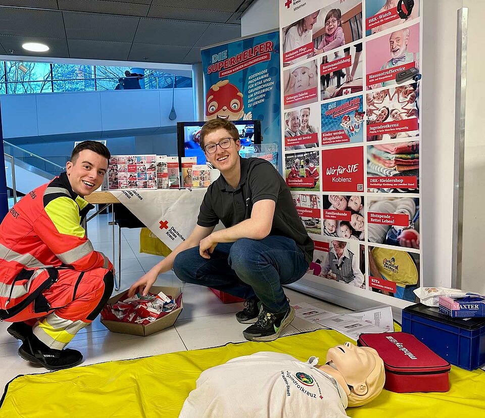 Notfallsanitäter und DRK-Mitarbeiter vor Logowand und Reanimations-Puppe